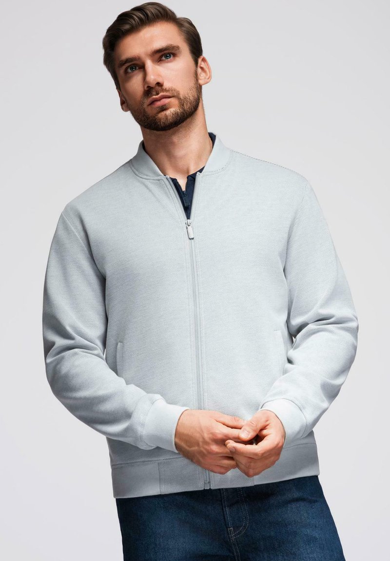 Uomo che indossa una giacca leggera grigia con zip e jeans scuri, posato con le mani incrociate, che guarda leggermente verso l'alto su uno sfondo semplice.