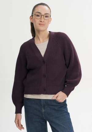 Woman with glasses wearing a dark purple buttoned cardigan, beige shirt, and blue jeans, standing with one hand in pocket on white background.