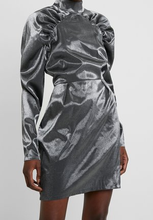 Srebrna, metaliczna mini sukienka z długim rękawem, wysokim kołnierzem i marszczonymi detalami na ramionach, noszona przez osobę o ciemnej karnacji na jednolitym tle.