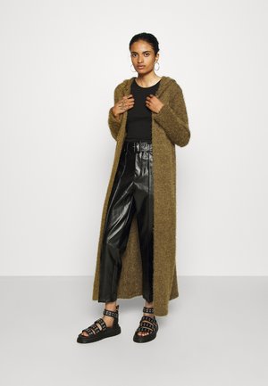 Long, fuzzy olive green cardigan over a black fitted top, paired with black leather trousers and strappy black sandals with accents.