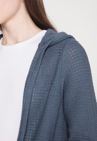 Lichtgewicht, gebreide hoodie met open voorkant in marineblauw met een gestructureerd patroon en een capuchon. Draagt als buitenlaag over een witte geribbelde longsleeve.