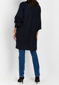 Pull en tricot bleu marine surdimensionné avec texture côtelée, manches longues, et fentes latérales, associé à un jean bleu ajusté et des talons noirs.