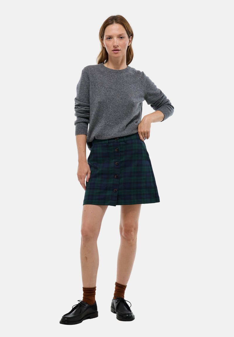 Jupe écossaise bleu marine et verte avec des boutons sur le devant, associée à un pull gris. Des chaussures noires à lacets et des chaussettes brunes complètent le look.