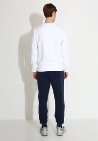 Bijela dukserica s rebrastim rukavima i donjim rubom, kombinirana s tamnoplavim trenirkama. Srebrne tenisice naglašavaju ležeran izgled. Pogled straga.