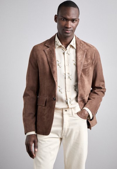 Blazer en daim marron avec revers crantés, deux poches avant et une fermeture à un bouton, superposé à une chemise à motif floral clair et un pantalon crème.