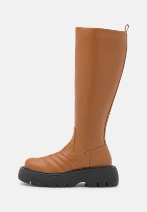 Höga bruna syntetläderstövlar med rund tå, ribbade detaljer och en tjock svart gummisula. Har en dragflik baktill.