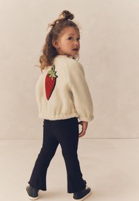 Hvid fluffy jakke med et jordbærmotiv på ryggen, kombineret med sorte vide bukser og sorte sneakers, mod en neutral baggrund.