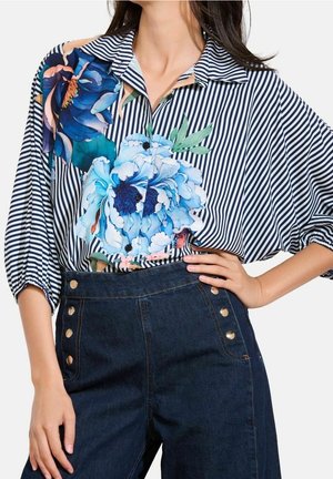Femme portant un chemisier rayé bleu et blanc avec de grands motifs floraux bleus et un pantalon en denim foncé taille haute avec des boutons dorés.