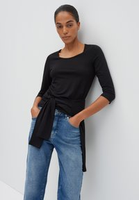 Zwarte langslijf top met een vierkante halslijn en geknoopt middel, gecombineerd met lichtblauwe wijde jeans, geplaatst tegen een effen achtergrond.