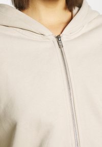 Gros plan d'une personne portant un sweat à capuche beige à fermeture éclair argentée, montrant la nuque et une partie des cheveux courts et bruns.