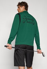 Grønn genser med svarte detaljer, med "Constantly Practicing" brodert på ryggen. Brukt med svarte shorts, holder en golfkølle.
