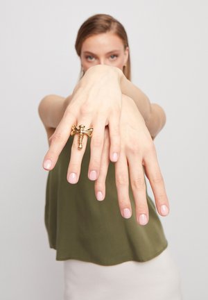 Bague en forme de libellule en or portée sur une main avec des ongles naturels. La bague présente des détails intriqués et une finition brillante. Le modèle porte un haut vert.