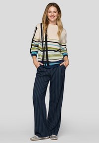 Mehrfarbiger gestreifter Pullover in Creme, Grün, Blau und Schwarz; kombiniert mit marineblauen, weit geschnittenen, gestreiften Hosen; Modell trägt silberne Schuhe.