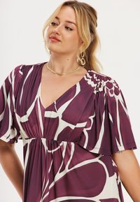 Lilla og hvidt mønstret kjole med korte flagreærmer, V-udskæring og rynket taljedetalje. Glat stof med flydende design.