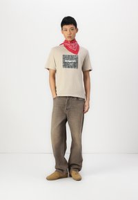 Camiseta beige con un gráfico cuadrado de patrón oscuro, combinada con unos vaqueros de mezclilla marrones de corte holgado y zapatos color beige. Una bandana roja está atada alrededor del cuello.