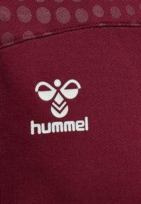 Abbigliamento sportivo color bordeaux con una texture liscia, caratterizzato da un logo "hummel" bianco e una grafica di un'ape, accentuato da un motivo a punti.