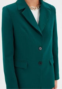 Blazer verde laguna con colletto a punta, due tasche frontali e tre bottoni neri. Realizzato in tessuto morbido, presenta un classico taglio sartoriale.
