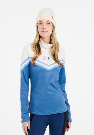 Blauwe longsleeve shirt met hoge kraag en zigzagpatroon in het wit, lichtblauw en roze. Gebreide crèmekleurige beanie op het hoofd van het model.