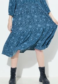 Sininen kukkamekko pitkillä hihoilla ja kootulla vyötäröllä, yhdistetty mustiin nauhakiilakorkoihin, esittelee virtaavan helman ja teksturoitua kangasta.