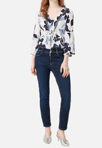 Mujer vistiendo una blusa blanca con estampado floral y mangas acampanadas, jeans ajustados oscuros y tacones plateados de punta, de pie contra un fondo sencillo.