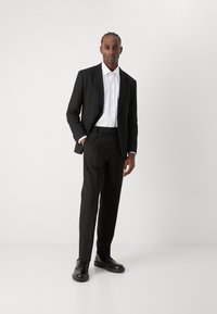 Schwarzer maßgeschneiderter Anzug mit einem weißen Hemd. Der Anzug hat einen klassischen Reverskragen und eine glatte Oberfläche. Schwarze Lederschuhe runden den Look ab.