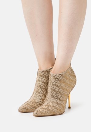 Bottines beiges à motifs avec talons aiguilles dorés, bouts pointus, portées sur jambes nues, sur fond blanc.