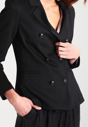 Blazer negro de doble botonadura con tejido texturizado, dos botones grandes, solapas afiladas y bolsillos laterales, creando una silueta ajustada.