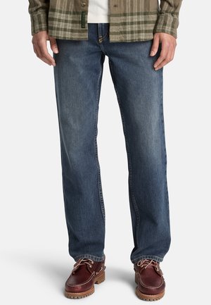 Mann trägt blaue Jeans mit geradem Bein, braune Leder-Schnürschuhe und ein grünes kariertes Hemd über einem weißen Top, steht vor weißem Hintergrund.