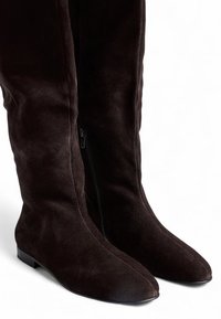 Bottes en daim marron jusqu'au genou avec une semelle plate et un bout pointu, dotées d'une fermeture éclair sur le côté intérieur pour un enfilage facile.