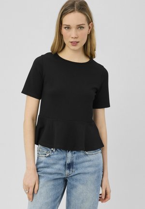 Jeune femme aux cheveux blonds lisses portant un haut péplum noir à manches courtes et un jean bleu clair, sur un fond uni.