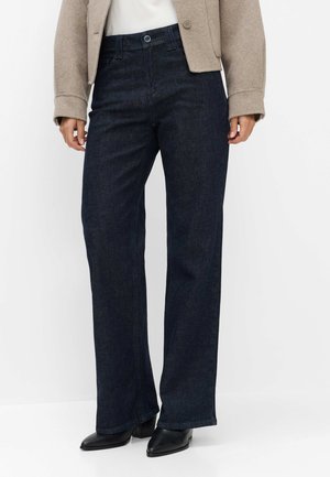 Person trägt dunkelblaue Jeans mit geradem Bein, beige Jacke, weißes Oberteil und schwarze spitze Pumps vor einfarbigem Hintergrund.