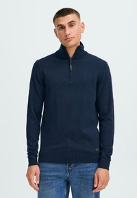 Pullover cu guler tip fermoar de culoare albastru marine, cu mâneci lungi, manșete și margine ribbrate, și un mic etichetă a brandului la talie. Textură moale și design ajustat.