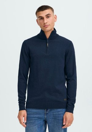 Mørkeblå zip-neck genser med lange ermer, ribbestrikket mansjetter og nederkant, samt et lite merke ved midjen. Myk tekstur og tettsittende design.