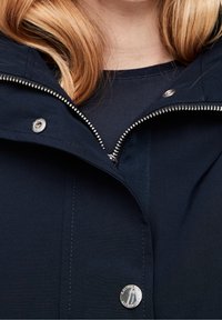 Chaqueta azul marino con cierre frontal, herrajes plateados, cuello alzado y una textura de tela suave con sutilezas en los detalles de costura.