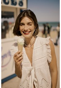 Svijetla pamučna bluzica s naboranim rukavima drži kornet svijetlozelene sladoledne kreme u vaflu, u pozadini plaže.
