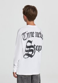 Biała koszulka z długim rękawem i okrągłym dekoltem, z czarnym, błyszczącym napisem na plecach: "Czas nigdy się nie zatrzymuje." Luźny krój, miękki materiał bawełnowy.