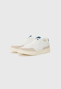 Zapatillas blancas con acentos de ante beige, que presentan una parte superior de cuero suave, puntera redonda, cordones planos y una etiqueta en el talón trasero azul. Suela texturizada.