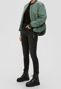 Grön bomberjacka med en glänsande yta, tillsammans med svarta tajta jeans och svarta platåsnörskor. Enkel, modern design med funktionella detaljer.