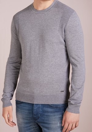 Man draagt een nauwsluitende grijze gebreide trui met geribbelde schouderaccenten en een blauwe jeans, staand tegen een effen achtergrond.