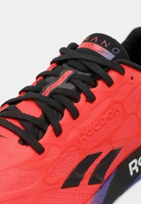 Shoe atlética vermelha com atacadores pretos, parte superior texturizada e marca Reebok. Apresenta detalhes em borracha e um colarinho acolchoado para conforto.