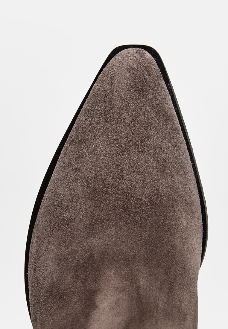 Scarpa in suede grigia con punta affusolata, caratterizzata da una texture liscia e un profilo elegante, con una sottile suola nera e accenti di design minimalisti.