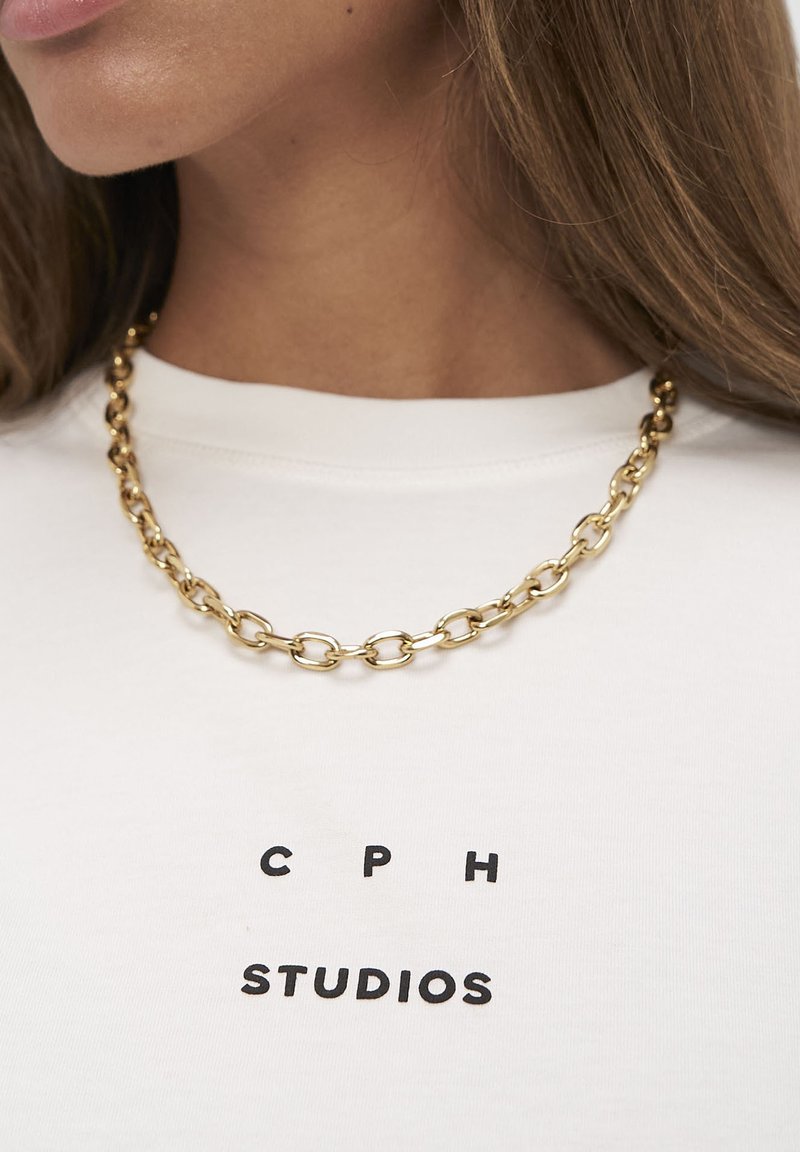 Guld kædehalskæde med ovale led, båret over en hvid skjorte med sort tekst, der lyder "CPH STUDIOS." Glat tekstur, metallisk glans.