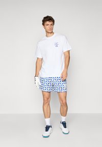Hvid bomulds t-shirt, blå grafisk design; mønstrede blå og hvide badeshorts; hvide sportsko; grå baggrund.