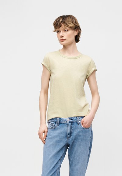 Νεαρός με κοντά μαλλιά που φοράει ένα ανοιχτό μπεζ μπλουζάκι με γυρισμένα μανίκια και μπλε τζιν, στέκεται με το ένα χέρι στην τσέπη.