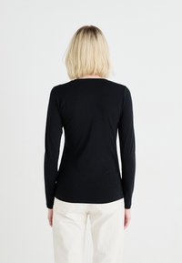 Blonde person wearing a fitted black long-sleeve shirt and cream pants, facing away against a plain white background.