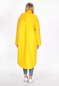 Sárga vízálló kabát kapucnival, hosszú ujjal és egyenes fazonnal, patentos mandzsettákkal, világoskék farmernadrág felett viselve, fehér cipővel.