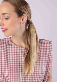 Lichtroze geruite jurk met wijd uitlopende mouwen; model heeft stijl, lang blond haar in een paardenstaart; draagt kleine gouden oorbellen met rode accenten.