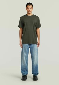 Donkergroene katoenen t-shirt met een ronde halslijn en korte mouwen, gecombineerd met lichtblauwe wijde jeans en zwarte schoenen.