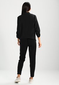 Schwarzes Langarmoberteil mit Kragen, leicht oversized geschnitten. Kombiniert mit schwarzen schmal geschnittenen Hosen und hellen Slippers.