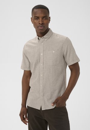 Mand iført lysebeige kortærmet skjorte med knapper og brystlomme samt mørke bukser, poserer med den ene hånd i lommen mod en ensfarvet baggrund.